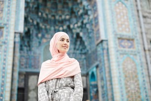 Contenido atractiva joven musulmana con hijab de pie contra la colorida mezquita con adornos y mirando hacia otro lado