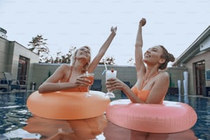 Happy ladies holding glasses of cocktails inside of rubber rings. Summer vacation concept
