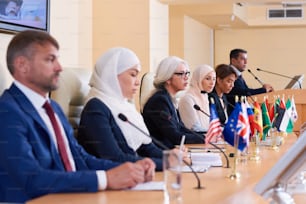 Reihe junger interkultureller Delegierter in formeller Kleidung, die bei der Konferenz am Tisch vor Mikrofonen sitzen