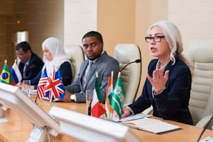 Mature well-dressed businesswoman explaining points of her report to audience while sitting among other delegates