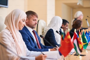 Young confident elegant businessman in formalwear sitting among his foreign partners and other speakers at conference