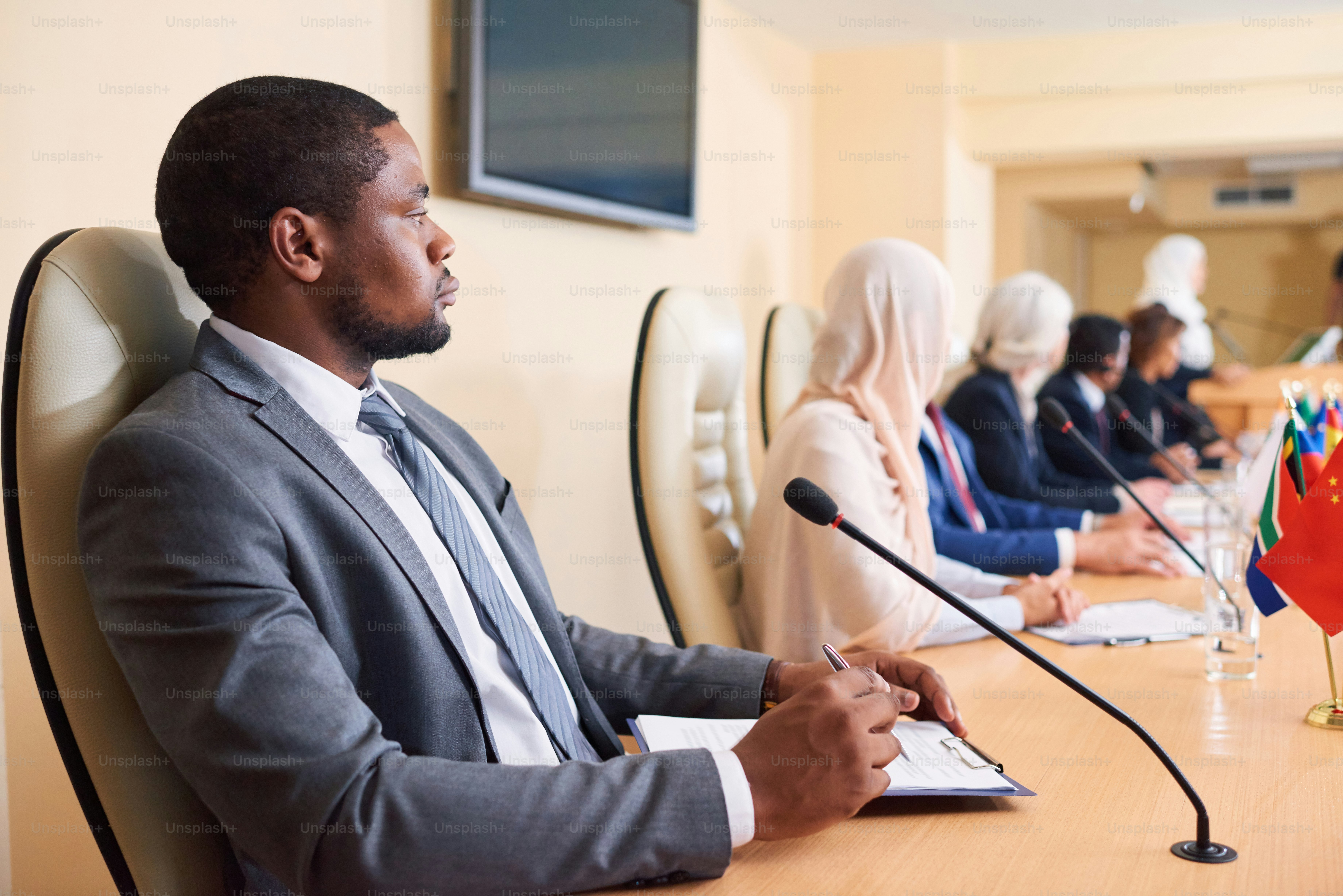 Selbstbewusster junger afroamerikanischer Delegierter in formeller Kleidung, der auf dem Hintergrund ausländischer Kollegen auf der Konferenz am Tisch sitzt