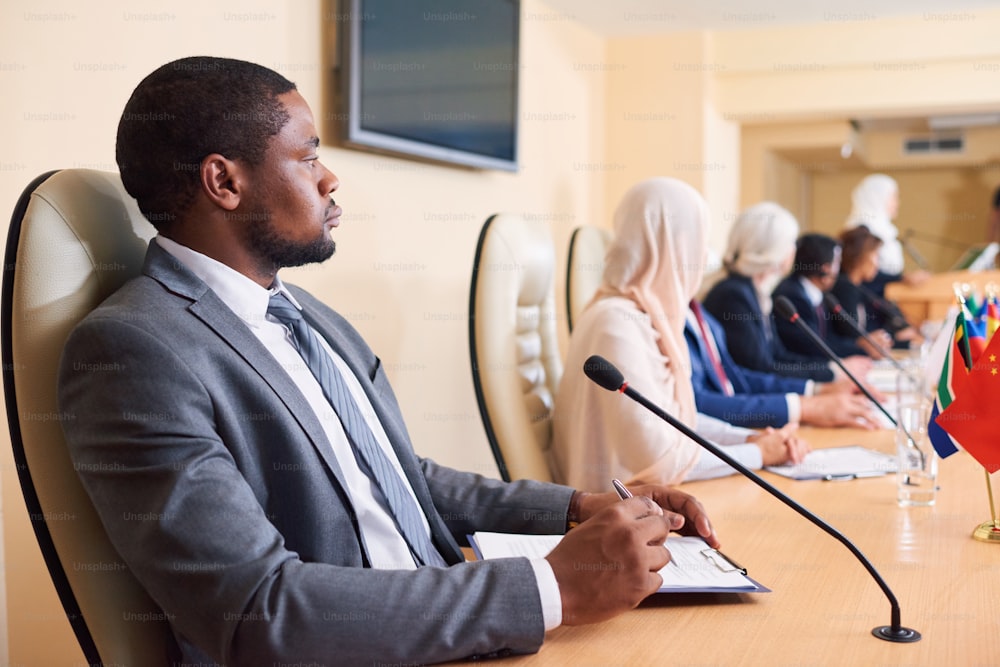 Confident young African-american delegate in formalwear sitting by table on background of foreign colleagues at conference