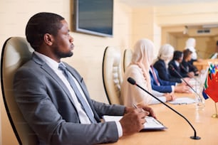 정장을 입은 자신감 넘치는 젊은 아프리카계 미국인 대표가 회의에서 외국 동료들의 배경에 테이블 옆에 앉아 있다