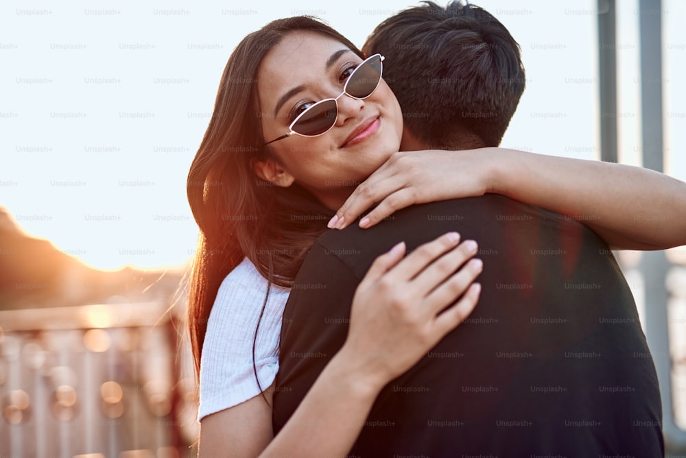 Liebes- und Date-Konzept. Junge fröhliche asiatische Dame mit Cat-Eye-Sonnenbrille umarmt ihren geliebten Mann
