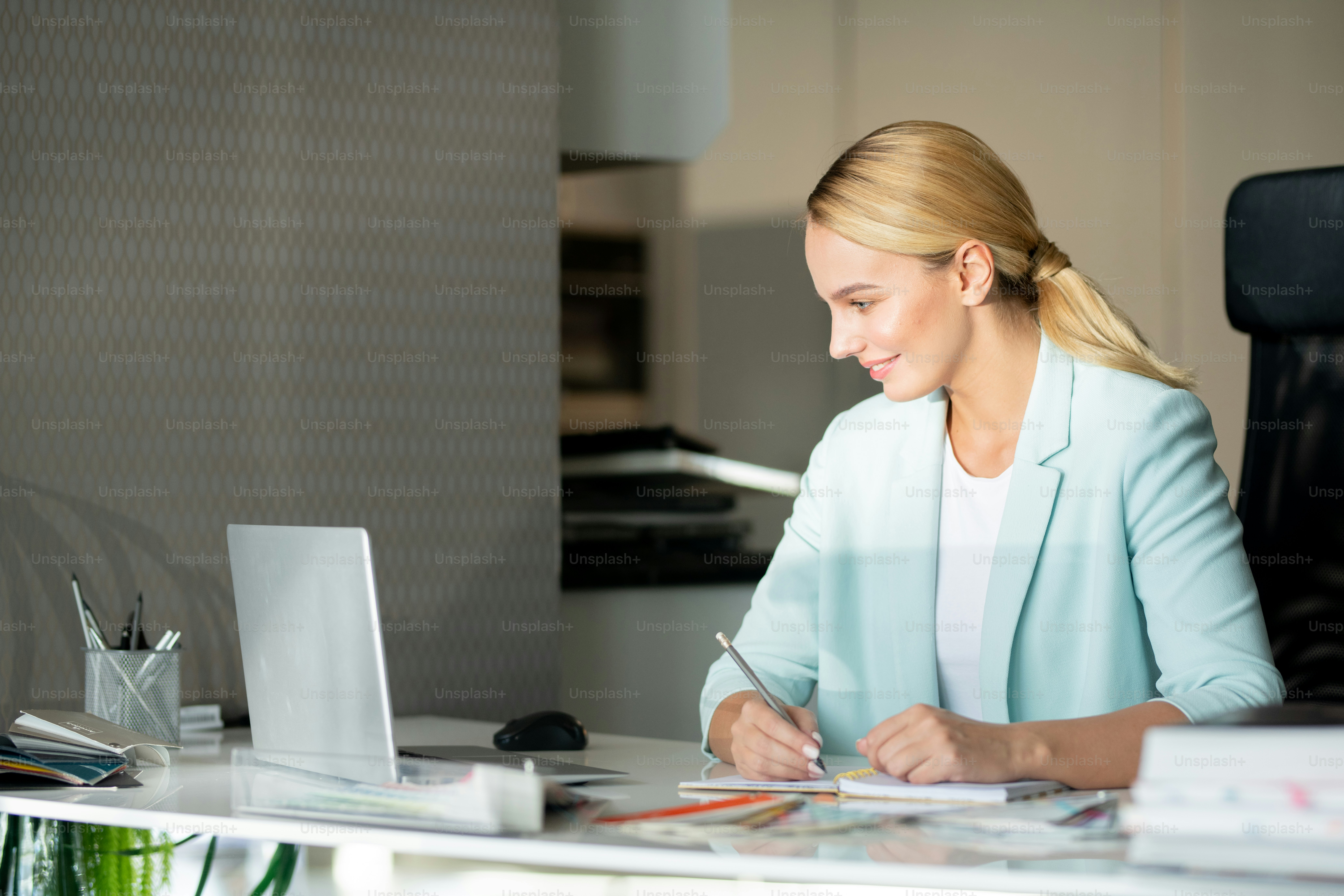 Young inspired designer sitting by desk in front of laptop, watching educational video and making notes
