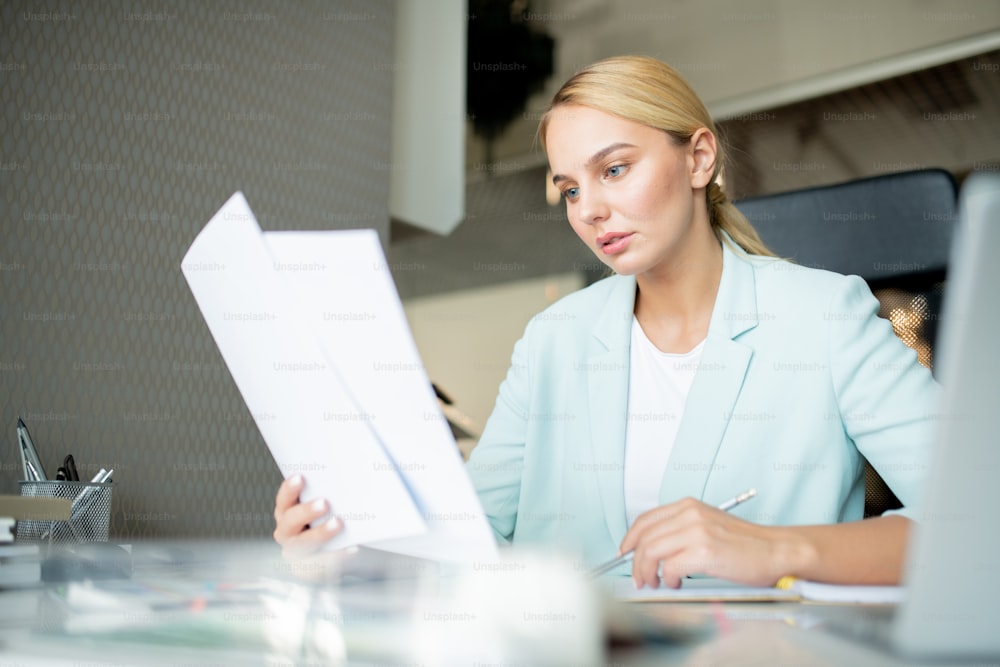 Young broker or businesswoman looking through text of new contract while sitting in office