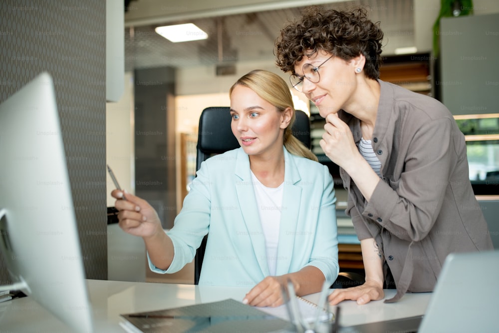 One of young creative designers pointing at computer screen while explaining her colleague some ideas