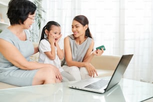 Asian little girl sitting together with mother and grandmother and looking at laptop with surprise they buying something with credit card online