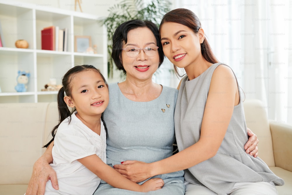 Portrait of happy Asian family generation mother daughter and grandmother embracing and smiling at camera while resting on couch