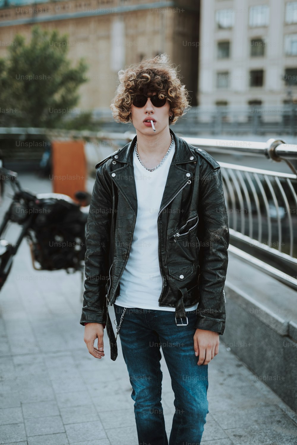 ストリートスタイルの革のジャケットを着た穏やかな若い巻き毛の男が歩き、喫煙する