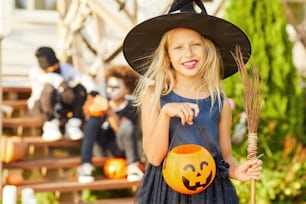 Cintura para cima retrato da menina bonito vestindo fantasia de Halloween olhando para a câmera enquanto posa ao ar livre segurando cesta de abóbora, espaço de cópia