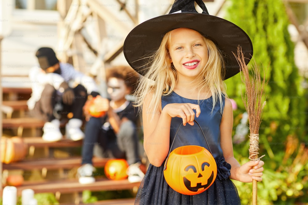 Retrato de cintura para arriba de una linda niña con disfraz de Halloween mirando a la cámara mientras posa al aire libre sosteniendo una canasta de calabazas, copiar espacio