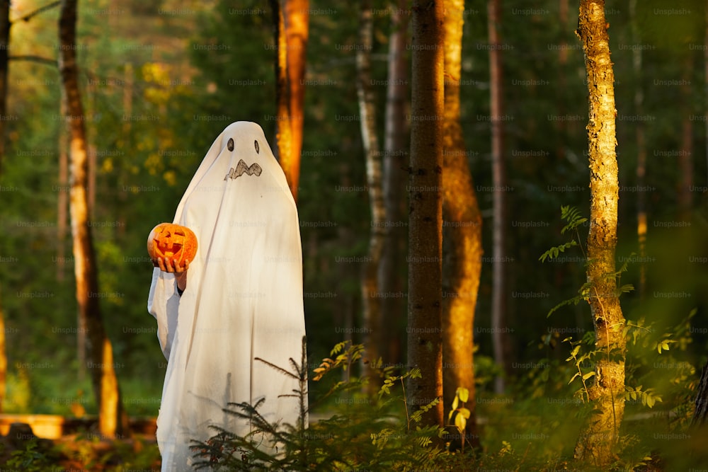 Ganzkörperporträt eines gruseligen Kindes, das als Geist verkleidet ist und Kürbis hält, der an Halloween im dunklen Wald steht, Kopierraum