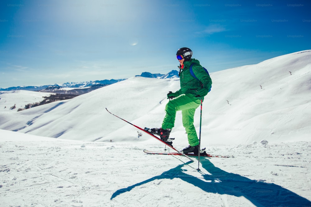 Esquiador em pé com uma perna levantada em uma pista de esqui em um dia ensolarado e olhando para algum lugar