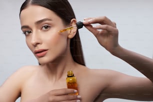 Close up portrait of beautiful naked lady with natural makeup holding glass dropper and bottle of orange liquid. Isolated on white background