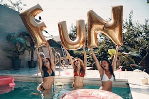 Attractive young women in swimwear smiling and lifting up balloons while standing in the pool outdoors