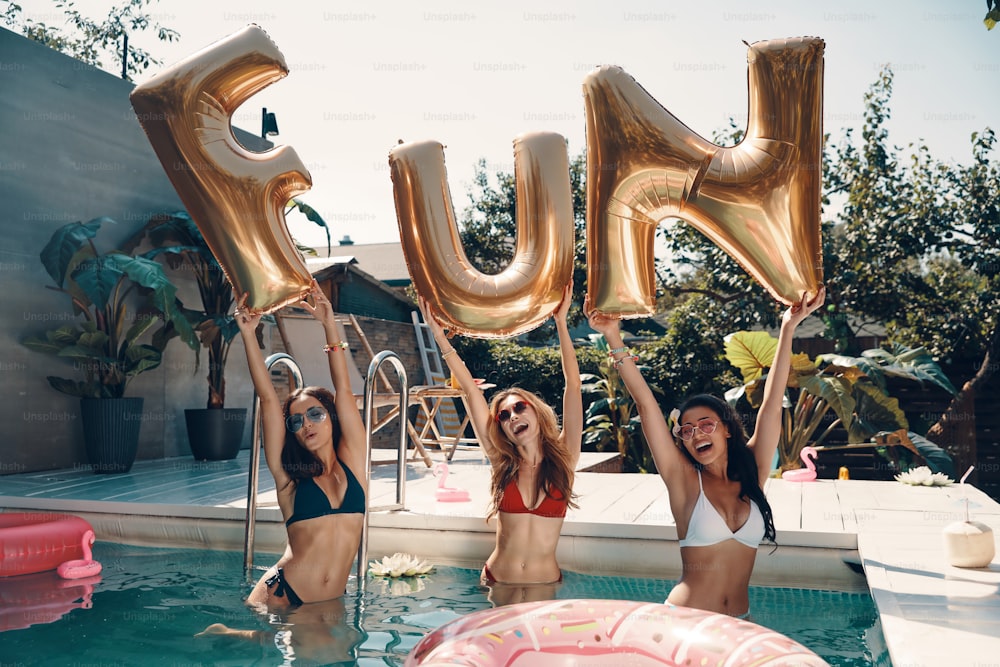 Mulheres jovens atraentes em trajes de banho sorrindo e levantando balões enquanto estão na piscina ao ar livre