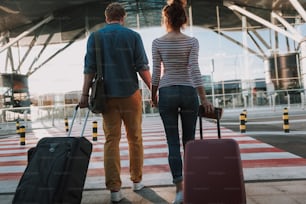 Full length back view portrait of young man and his charming girlfriend walking and carrying their trolley bags