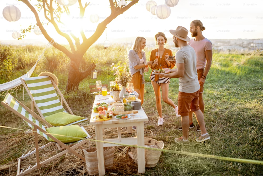 Gruppe von jungen und glücklichen Freunden, die sich bei einem festlichen Picknick im wunderschön dekorierten Garten mit Mittagstisch bei Sonnenuntergang amüsieren