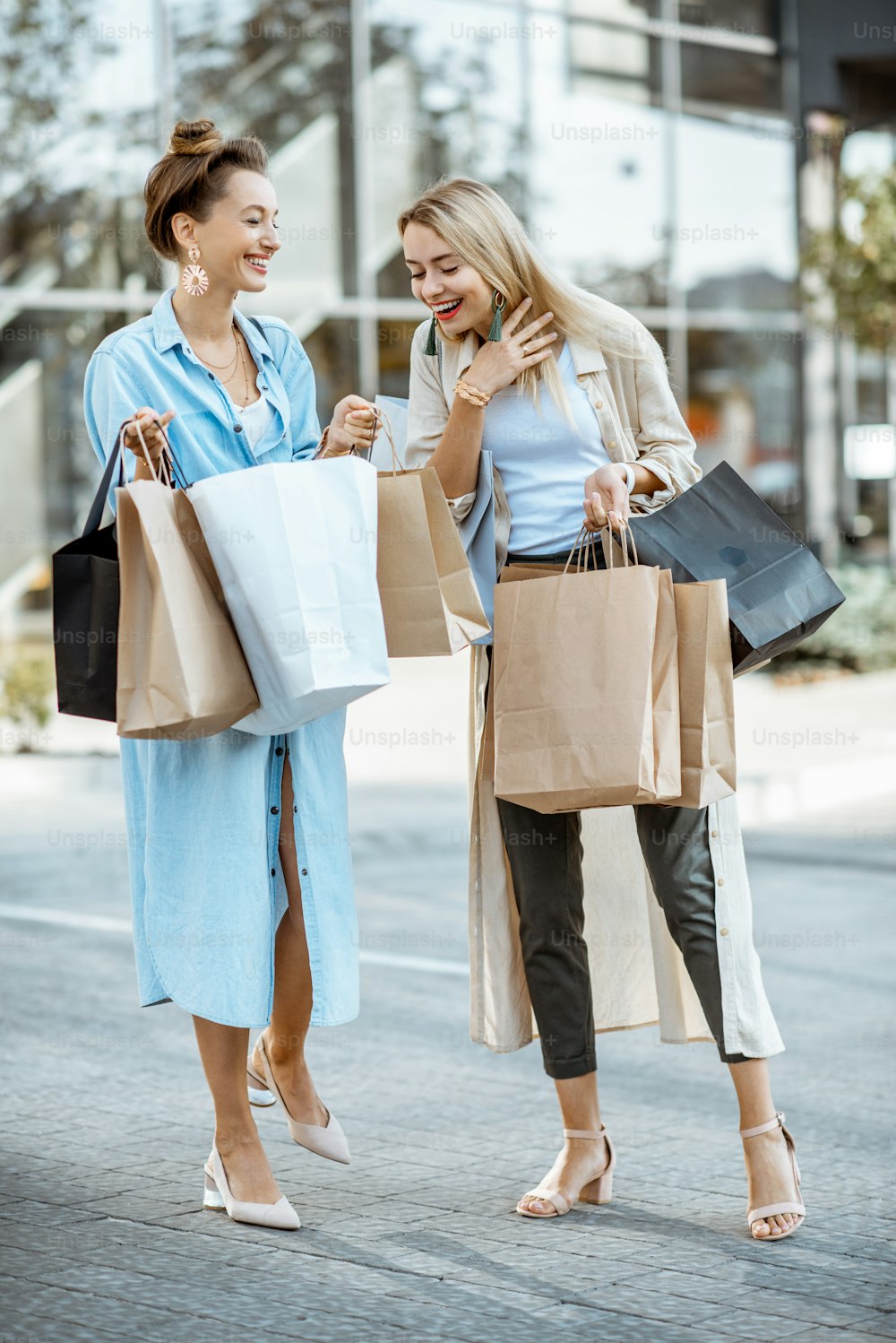 買い物に満足している幸せな2人のガールフレンドは、屋外のショッピングモールの前で買い物袋を持って一緒に立っています