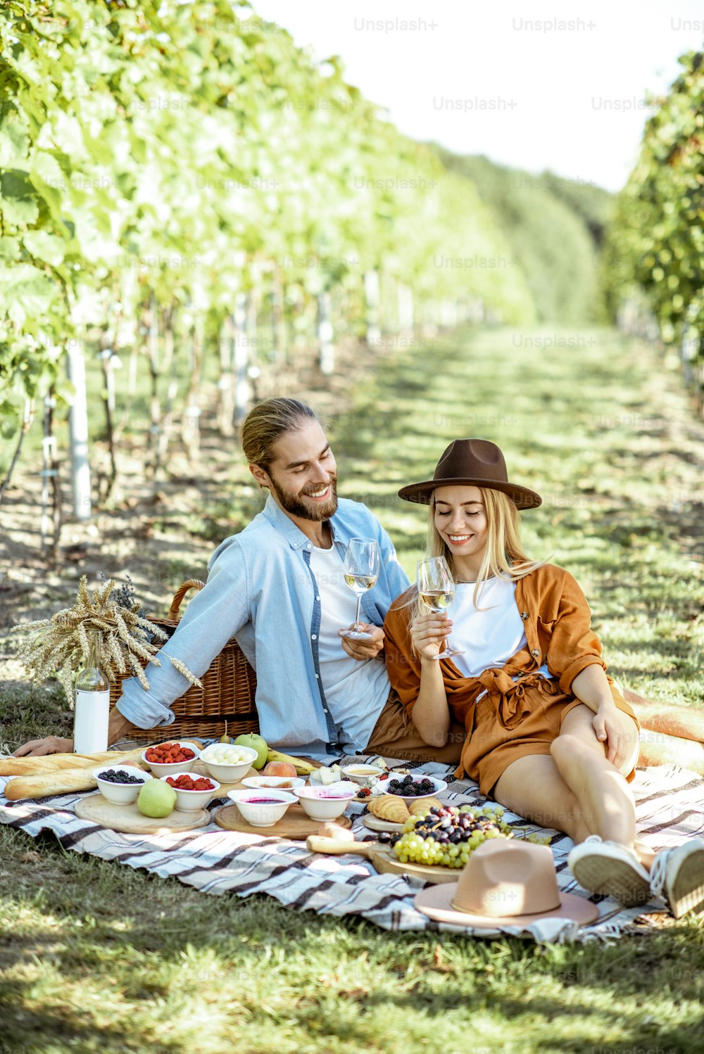 Hermosa pareja que tiene un desayuno romántico con mucha comida sabrosa y vino, sentados juntos en la manta de picnic en el viñedo en una mañana soleada