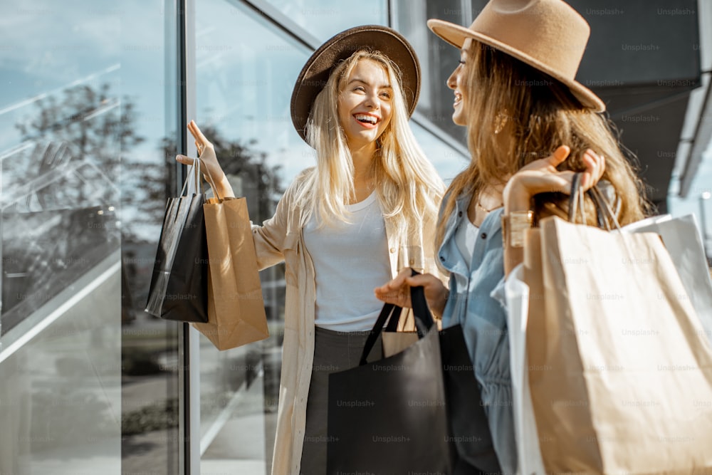 쇼핑몰 근처에 쇼핑백을 들고 서 있는 동안 상점 창을 바라보는 두 명의 행복한 여자 친구