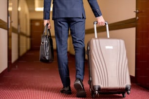 Rückansicht eines jungen reisenden Geschäftsmannes im Anzug, der eine schwarze Lederhandtasche trägt und einen Koffer zieht, während er sich entlang des Korridors bewegt