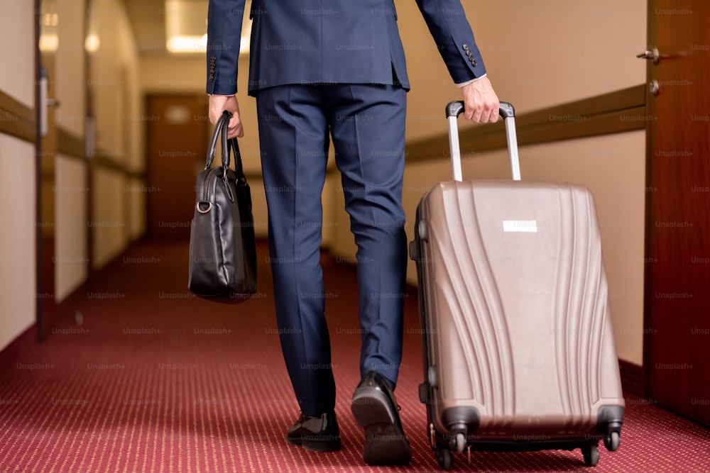 Rear view of young traveling businessman in suit carrying black leather handbag and pulling suitcase while moving along corridor