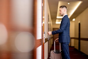 Giovane uomo d'affari con il bagaglio che tiene la carta di plastica dalla porta mentre entra nella stanza dopo l'arrivo in hotel