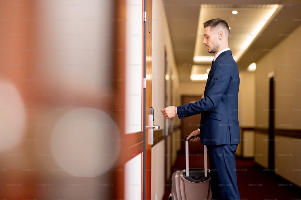 Young businessman with baggage holding plastic card by door while going to enter the room after arriving in hotel