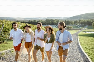 Eine Gruppe junger Freunde, die lässig gekleidet sind, hängen zusammen ab und gehen an einem sonnigen Tag mit Weingläsern auf dem Weinberg spazieren
