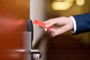 Mano de un hombre de negocios moderno sosteniendo una tarjeta de plástico roja junto a la puerta de madera cerrada de la habitación del hotel para entrar