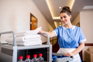 Young smiling chamber maid taking white clean towel from top of stack while pushing cart with hygiene stuff
