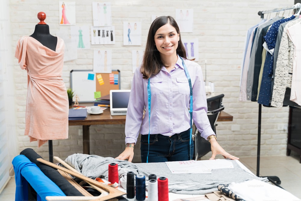 Portrait of smiling young beautiful fashion designer standing at workbench in atelier
