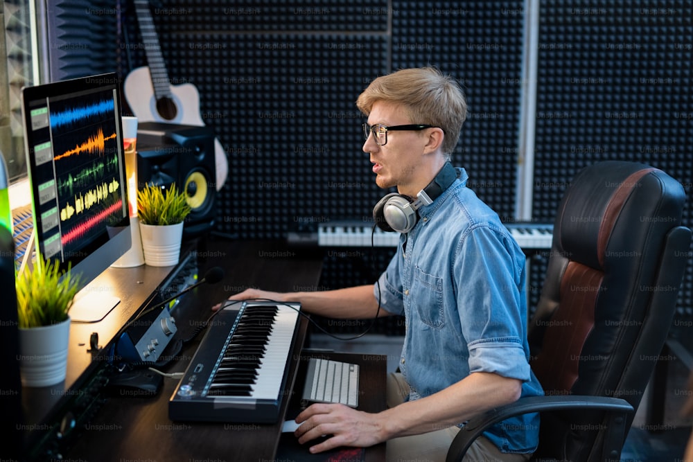 Giovane deejay maschio in camicia di jeans che guarda i traclis di mixaggio del suono sullo schermo del computer mentre è seduto alla scrivania davanti al monitor