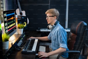 Giovane deejay maschio in camicia di jeans che guarda i traclis di mixaggio del suono sullo schermo del computer mentre è seduto alla scrivania davanti al monitor