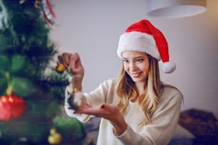Beautiful caucasian young woman dressed in sweater and with Santa's hat on head decorating christmas tree while standing in living room.