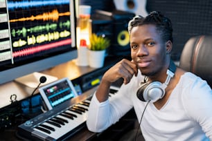 Happy young smiling mixed-race man with headphones on neck sitting by workplace with piano keyboard and computer monitor