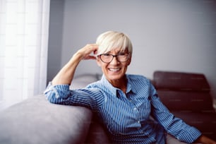 Splendida donna anziana sorridente vestita con camicia a righe e con occhiali da vista in posa e guardando la macchina fotografica.