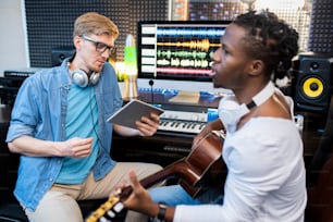 アフリカ民族の若い歌手またはミュージシャンがギターを弾き、同僚がスタジオで曲を録音しながら歌う