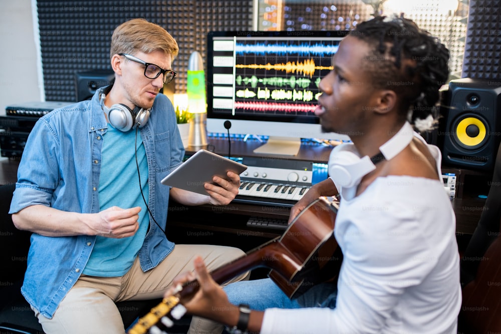 Young singer or musician of African ethnicity playing the guitar and singing while his colleague recording the song in studio