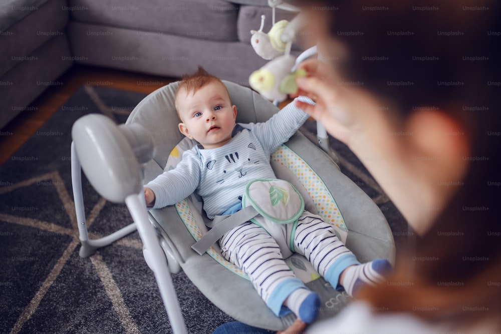 Adorable baby boy lying in bouncer an looking at his mother. Mother entertaining her loving son. Living room interior.