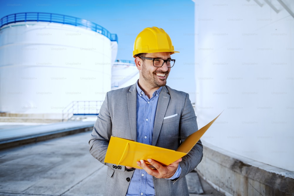 Handsome caucasian businessman in suit standing outdoors and holding folder. In background are oil storage tanks.