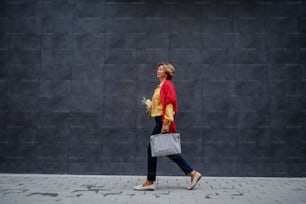 Vista laterale dell'affascinante donna anziana alla moda caucasica che trasporta borsa e generi alimentari mentre passa dal muro grigio.