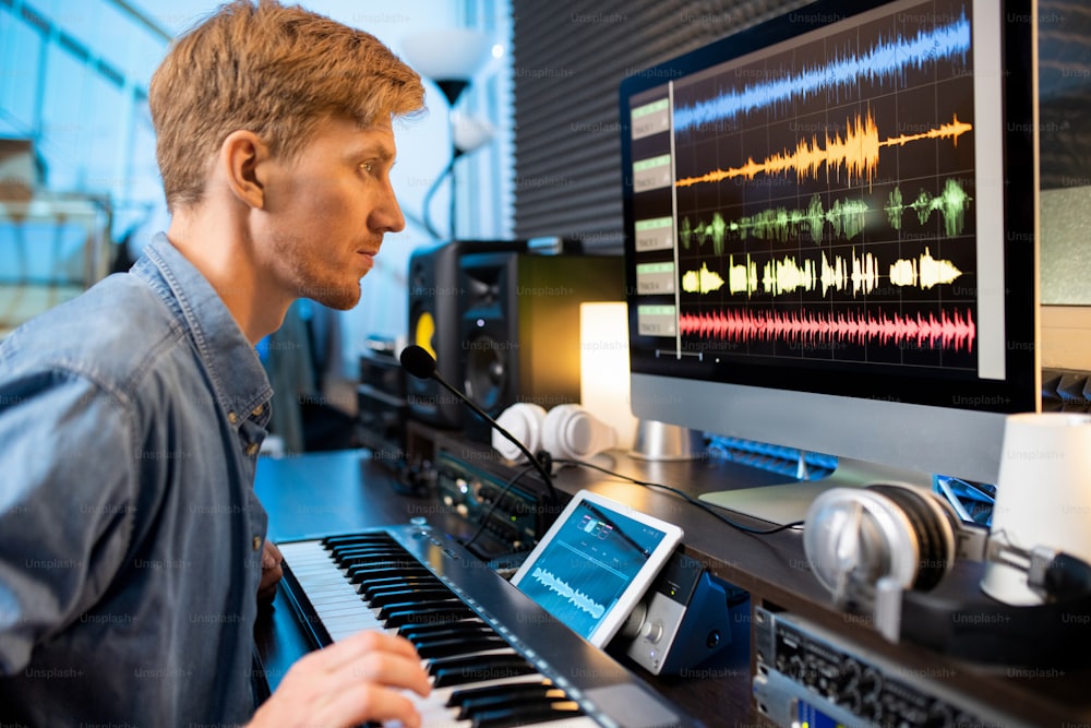 Homem sério tocando uma das teclas do pianoboard enquanto olha para as formas de onda do som na tela do computador na frente