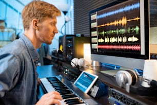 Homem sério tocando uma das teclas do pianoboard enquanto olha para as formas de onda do som na tela do computador na frente
