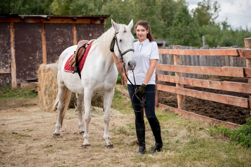 Joven mujer activa en jeans, polo y botas mirándote mientras te relajas con un caballo de carreras blanco de pura raza en un entorno rural