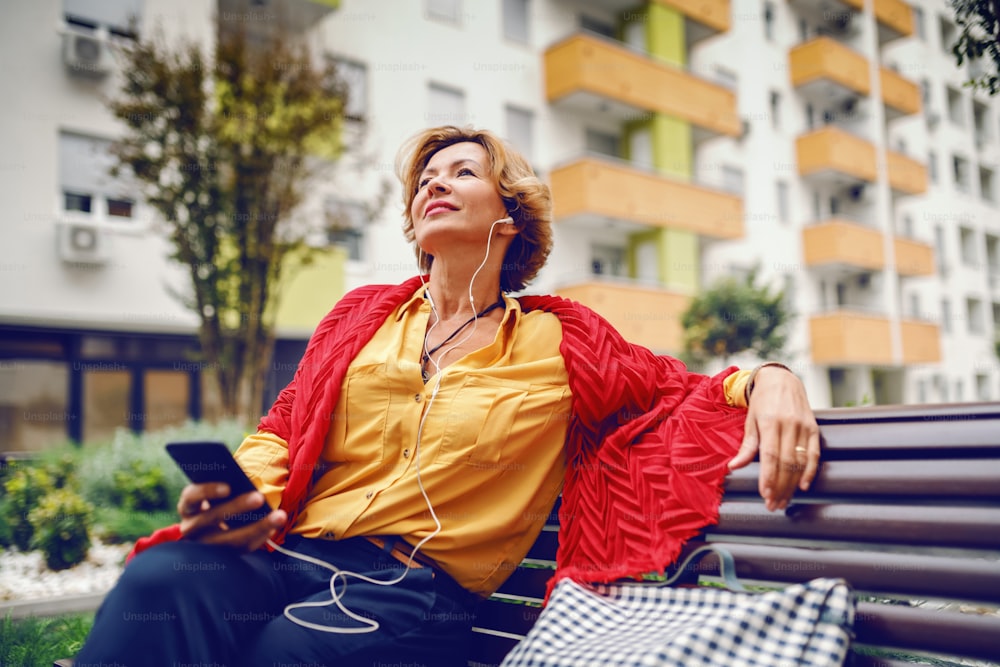 Beautiful Caucasian blonde fashionable senior woman sitting on bench in park, holding smart phone and listening music. In background are buildings.