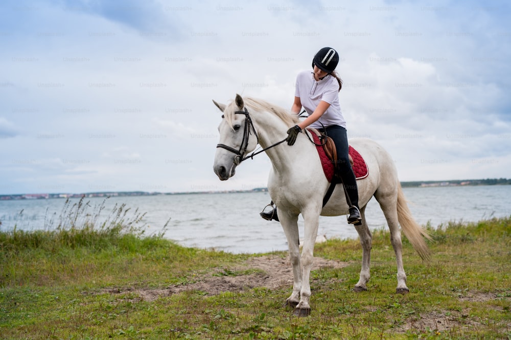 Junge aktive Frau in Reiterkleidung, die sich am Flussufer entlang bewegt, während sie während des Trainings auf dem Rücken eines weißen Rennpferdes sitzt
